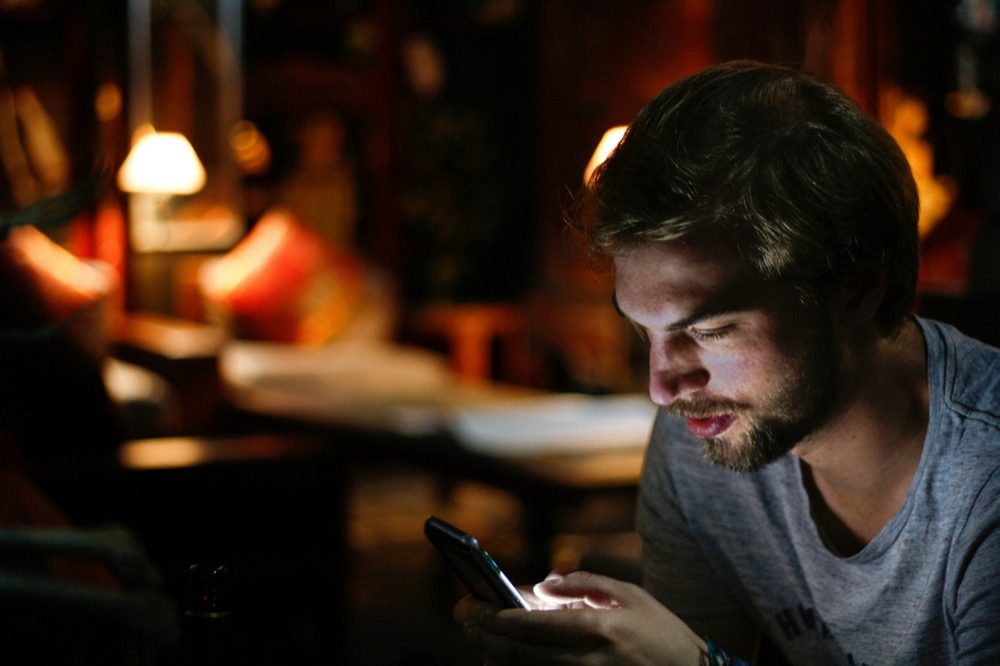 Homem de barba olhando para o celular, com o rosto iluminado pela tela, em um ambiente com luz baixa